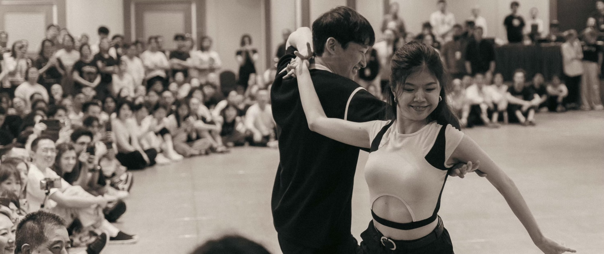 Two dancers perform together while a seated audience watches around the dance floor